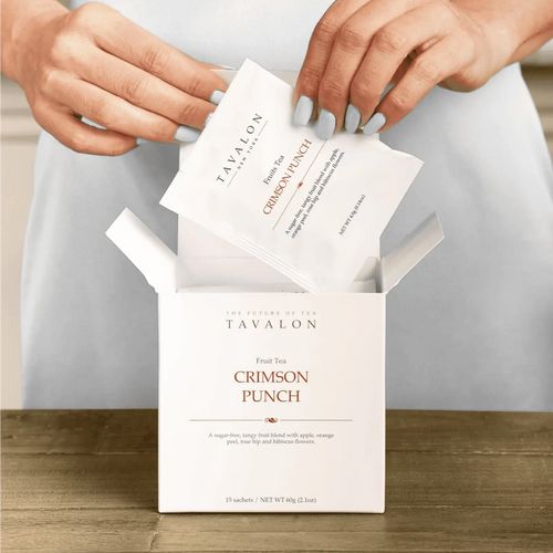 Tavalon Crimson Punch tea bags being taken out of a box on a wooden surface.