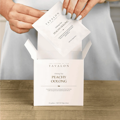 Tavalon Peachy Oolong tea packaging with a hand opening it on a wooden surface.